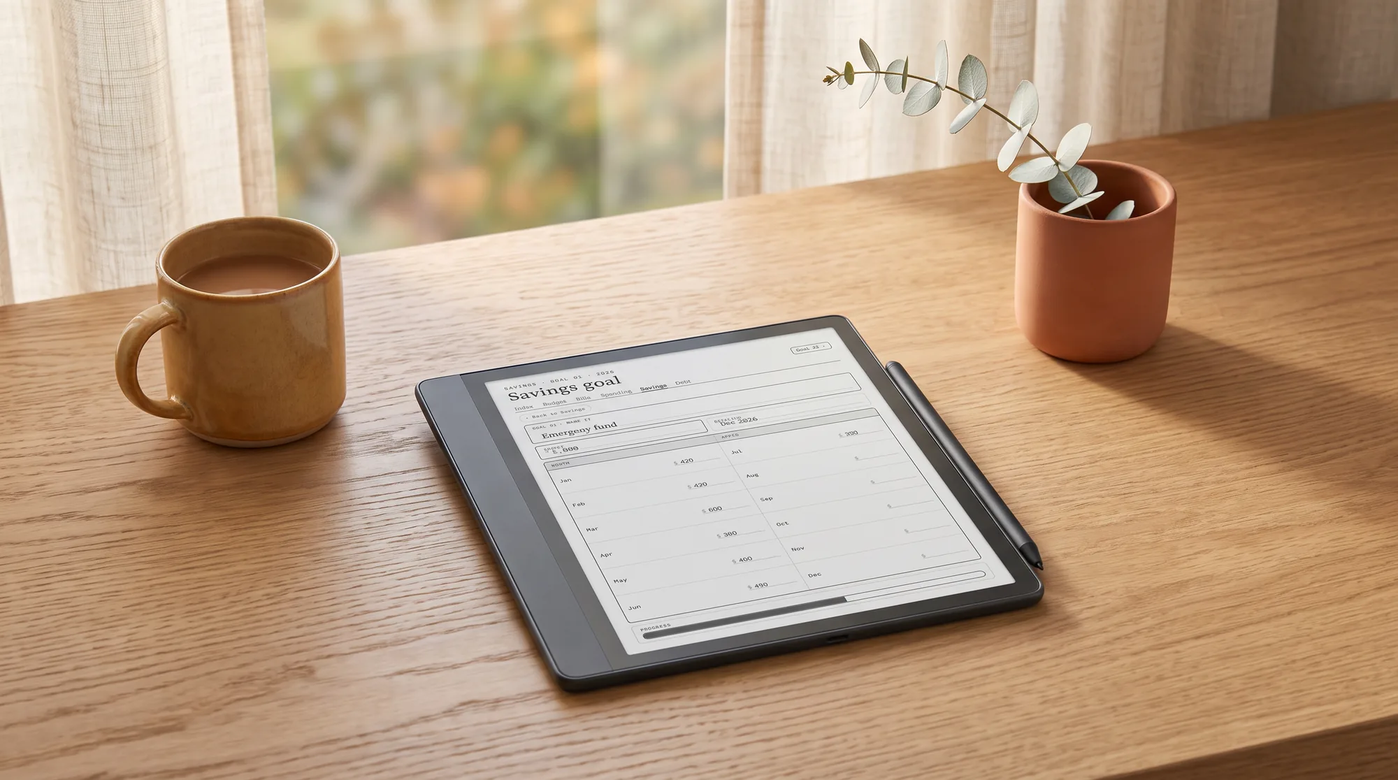 A Kindle Scribe showing a savings goal tracker, laid on a desk with a eucalyptus sprig and a mug of coffee